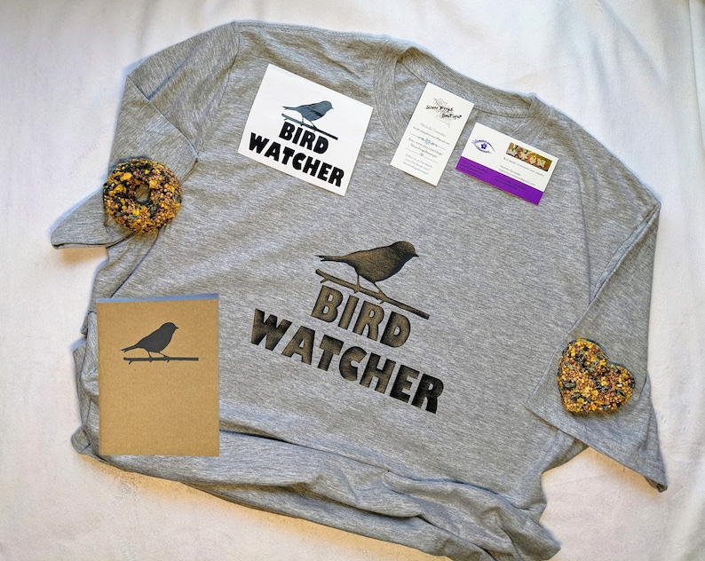 May include: A grey t-shirt with the text "Bird Watcher" and a silhouette of a bird. The shirt is packaged with a card, two stickers, and two small bags of birdseed.