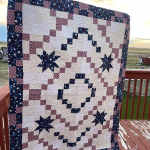 May include: A quilt with a patchwork design featuring a white background, pink and blue squares, and a central motif of stars. The quilt is bordered with a blue and white fabric with stars. The quilt is hanging on a wooden railing.