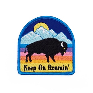 May include: An embroidered patch featuring a black buffalo silhouette against a colourful sunset backdrop. The patch depicts a blue sky with a yellow sun, white mountains, and a rainbow-coloured horizon. The text "Keep On Roamin'" is embroidered in yellow below the buffalo.