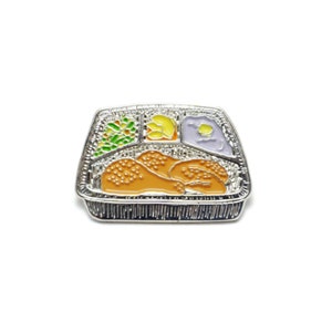 Puede incluir: Un pin de esmalte plateado con forma de bandeja de cena televisiva clásica. La bandeja está dividida en secciones que contienen pollo frito, puré de patatas, verduras y un acompañamiento amarillo. El pin está sobre un fondo blanco.