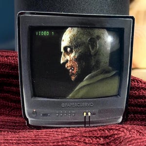 May include: A sticker featuring a retro television screen displaying a zombie's face. The screen reads "VIDEO 1." The sticker has a black border and the text "@PAPERCURVO" is visible. The background is a dark red fabric.