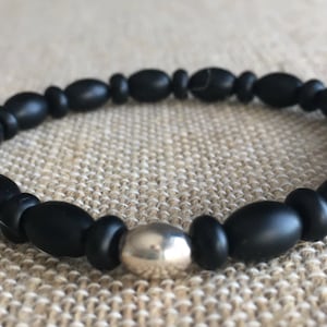 May include: A black beaded bracelet with a silver-colored central bead. The bracelet features oval-shaped beads strung together, creating a simple yet elegant design. The beads have a matte finish, and the bracelet is displayed on a neutral-toned surface.