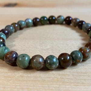 May include: A beaded bracelet featuring round, polished beads in shades of green, brown, and tan. The bracelet is strung on an elastic cord and rests on a light-colored wooden surface. This accessory is suitable for everyday wear.
