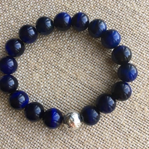 May include: A beaded bracelet featuring large, round, polished beads in shades of deep blue and a single silver-colored bead. The bracelet is displayed on a neutral-toned, textured surface.