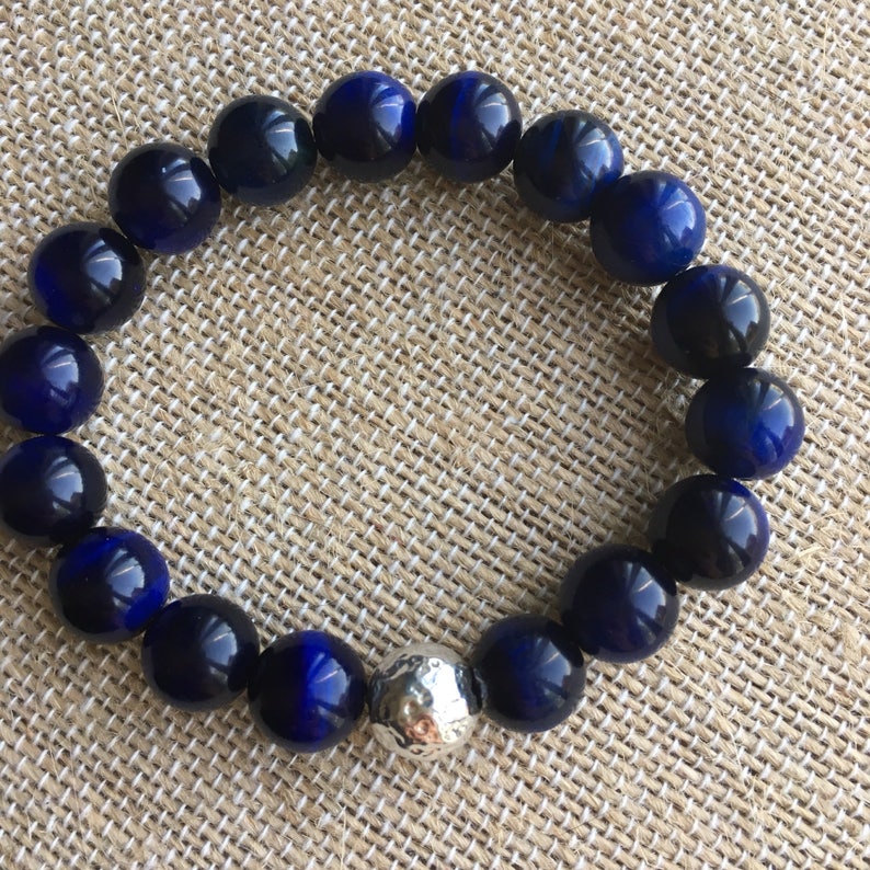 May include: A beaded bracelet featuring large, round, polished beads in shades of deep blue and a single silver-colored bead. The bracelet is displayed on a neutral-toned, textured surface.