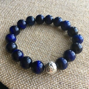 May include: A beaded bracelet featuring dark blue and black spherical beads. A single silver-colored, textured bead is interspersed among the dark blue beads. The bracelet is displayed on a tan, textured surface.