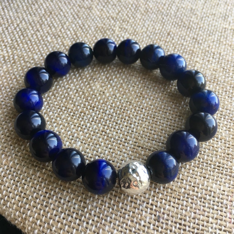 May include: A beaded bracelet featuring dark blue and black spherical beads. A single silver-colored, textured bead is interspersed among the dark blue beads. The bracelet is displayed on a tan, textured surface.