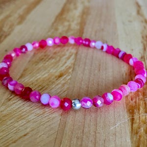 May include: A pink beaded bracelet featuring a mix of pink and white beads. The bracelet includes a silver-coloured bead at its centre. The faceted, round beads are strung together on elastic. The bracelet is displayed on a wooden surface.