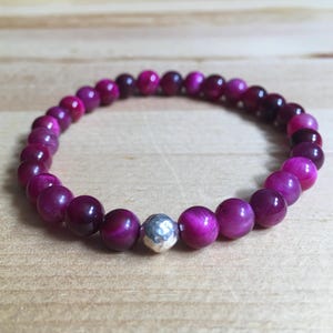 May include: A beaded bracelet featuring round, polished beads in shades of pink and purple. A single, silver-colored, faceted bead is interspersed among the other beads. The bracelet is displayed on a light brown wooden surface.
