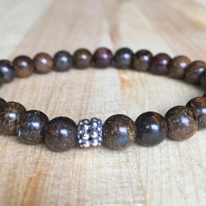 Peut inclure: Un bracelet de perles rondes marron foncé et dorées. Une perle d'espacement argentée est centrée sur le bracelet. Le bracelet est présenté sur une surface en bois brun clair.
