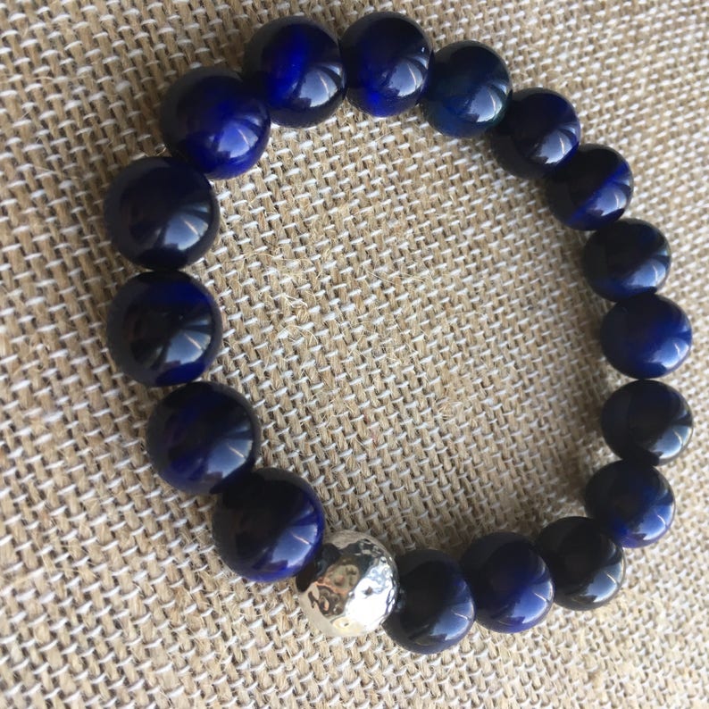 May include: A dark blue beaded bracelet with a silver accent bead. The bracelet features large, round, polished beads in a deep navy color. The silver bead has a hammered texture. The bracelet is displayed on a neutral-toned woven surface.