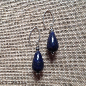 May include: A pair of earrings with deep blue lapis lazuli teardrop pendants. The earrings have silver-coloured, curved ear wires. The lapis lazuli stones are polished and hang against a textured, neutral background.