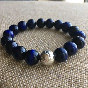 May include: A beaded bracelet featuring dark blue and silver beads. The bracelet is made of round, polished dark blue beads with a single silver, textured bead. The bracelet is displayed on a neutral-colored surface.