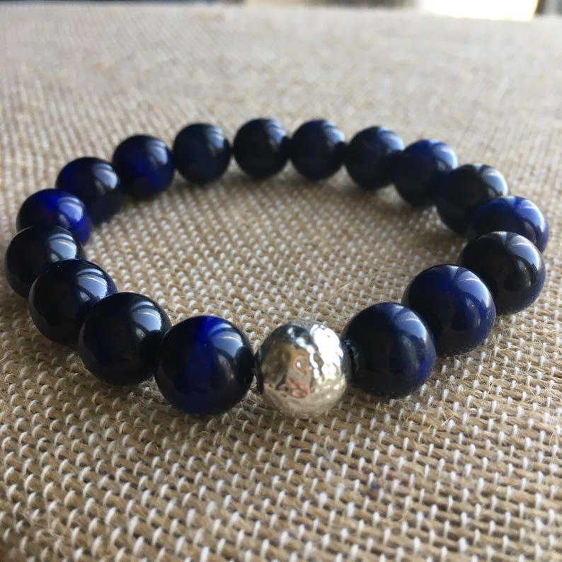 May include: A beaded bracelet featuring dark blue and silver beads. The bracelet is made of round, polished dark blue beads with a single silver, textured bead. The bracelet is displayed on a neutral-colored surface.