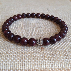May include: A dark red gemstone bracelet with a silver bead accent. The bracelet is made of round beads and is approximately 7 inches in length.