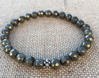 May include: A pyrite beaded bracelet. The bracelet features faceted, metallic grey beads strung together with a small silver-coloured accent bead. The bracelet is displayed on a neutral-coloured, textured surface.