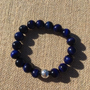 May include: A dark blue beaded bracelet with a silver accent bead. The bracelet is made of round, polished beads with a reflective quality. The beads are strung together on an elastic band. The bracelet is displayed on a textured, tan surface.