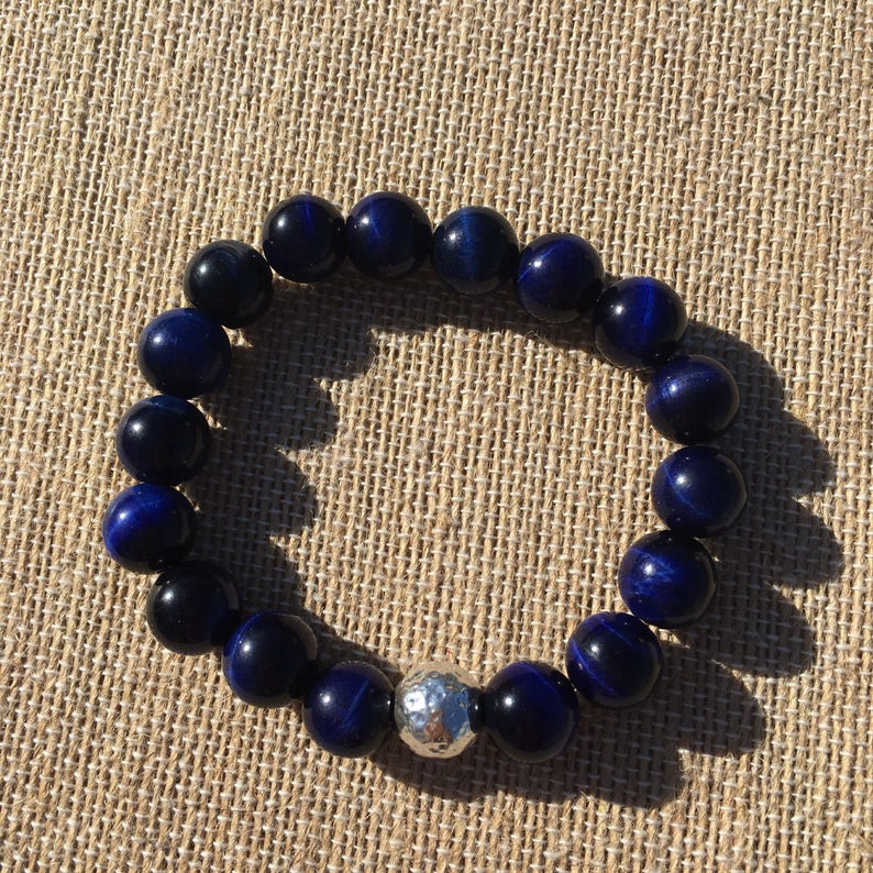 May include: A dark blue beaded bracelet with a silver accent bead. The bracelet is made of round, polished beads with a reflective quality. The beads are strung together on an elastic band. The bracelet is displayed on a textured, tan surface.