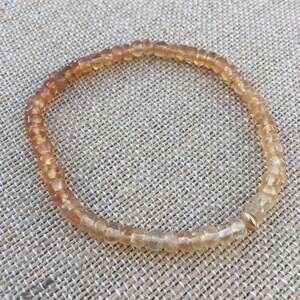 May include: A light brown beaded bracelet with a gold accent. The bracelet is made of small, round beads and is strung on a thin cord.