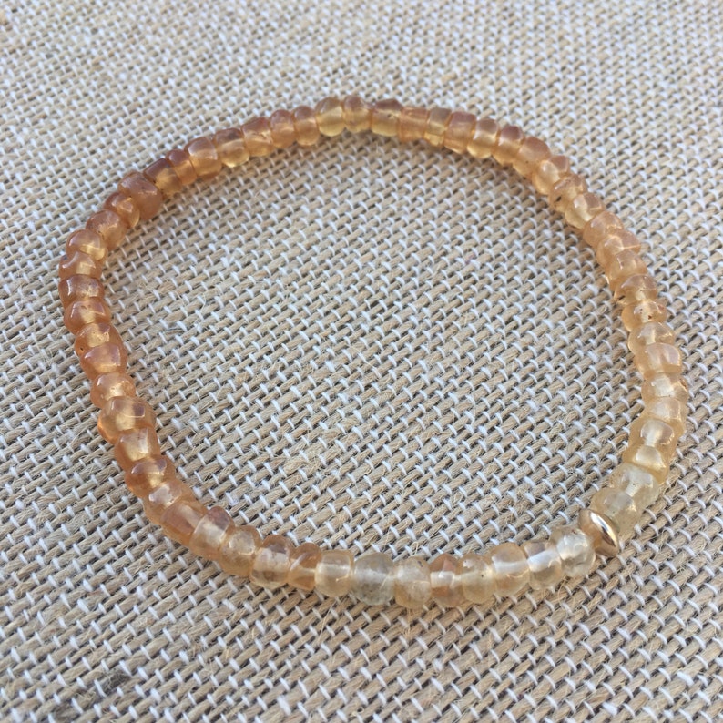 May include: A light brown beaded bracelet with a gold accent. The bracelet is made of small, round beads and is strung on a thin cord.
