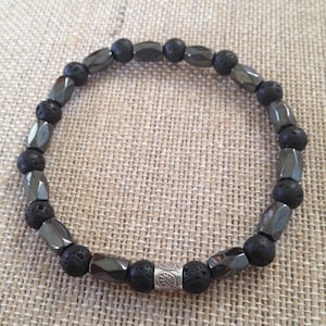May include: A black beaded bracelet with a mix of faceted hematite and porous lava rock beads. The bracelet includes a silver-toned spacer bead with a leaf design. The bracelet is displayed on a neutral, textured surface.
