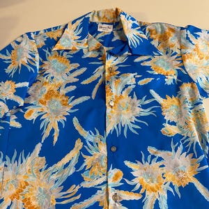 May include: A blue short-sleeved shirt with a floral print. The shirt has a button-up front and a pocket on the left side. The label on the shirt reads "Hawaii Nei".