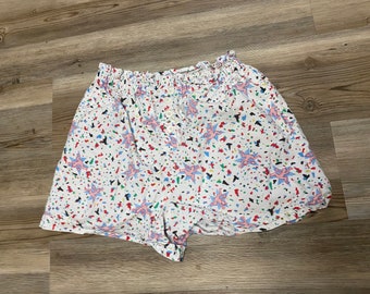 May include: White cotton shorts with a colourful confetti print and pink cartoon figures. The shorts have an elastic waistband.