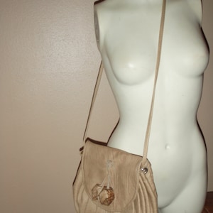 Puede incluir: Un bolso bandolera de cuero marrón claro con cierre de solapa y dos botones de madera. El bolso está en un maniquí blanco.