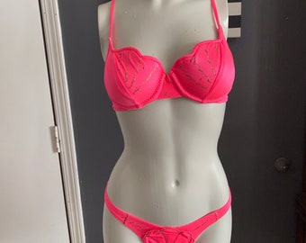 Könnte beinhalten: Ein pinkfarbener Bikini mit goldenen Akzenten. Das Oberteil hat einen gewellten Saum und das Unterteil ein herzförmiges Design.