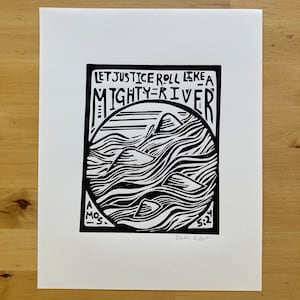May include: Black and white art print with the text "LET JUSTICE ROLL LIKE A MIGHTY RIVER" above a circular image of mountains and water. The print includes the text "AMOS 5:24" and the artist's signature.