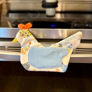 May include: A fabric chicken-shaped oven towel featuring a white background with a multi-colored floral and chicken pattern, a bright orange comb, a yellow beak, and a solid light blue wing-shaped patch centered on the body.