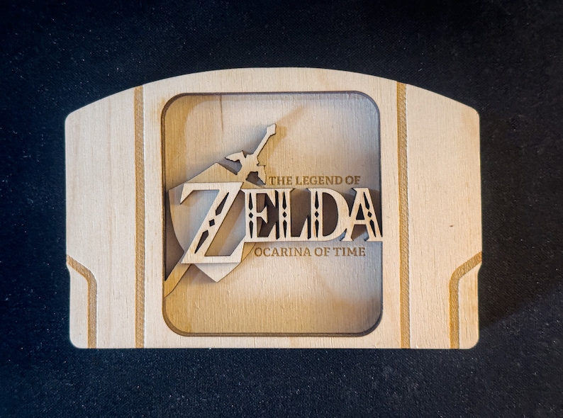 May include: A wooden Nintendo 64 game cartridge case with a laser-cut design featuring the "The Legend of Zelda: Ocarina of Time" logo.