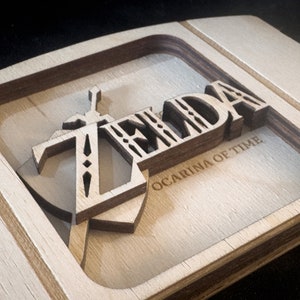 May include: A wooden cutout of the Legend of Zelda: Ocarina of Time logo. The logo is made of multiple layers of wood and features the iconic Triforce symbol and the words "ZELDA" and "OCARINA OF TIME".