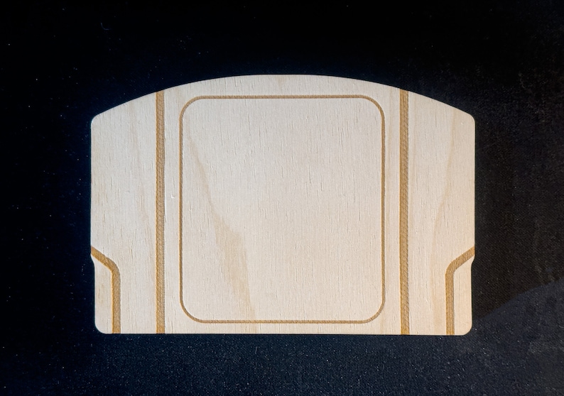 May include: A blank wooden Nintendo 64 game cartridge case with a light brown wood grain. The case has a rectangular opening in the centre and is outlined with a thin brown line.