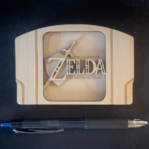 May include: A wooden Nintendo 64 game cartridge case featuring a laser-cut design of the "The Legend of Zelda: Ocarina of Time" logo.