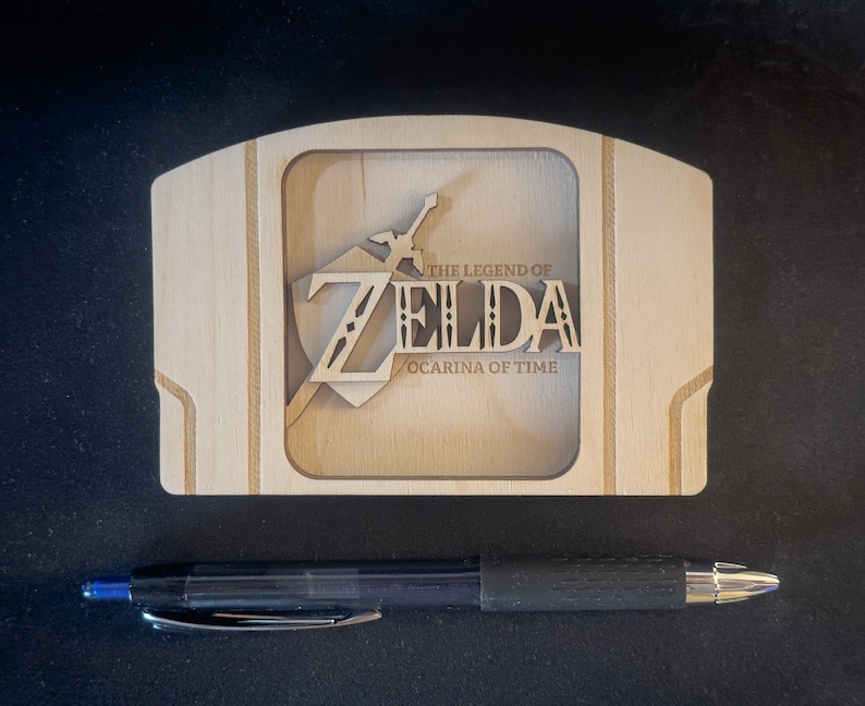 May include: A wooden Nintendo 64 game cartridge case featuring a laser-cut design of the "The Legend of Zelda: Ocarina of Time" logo.