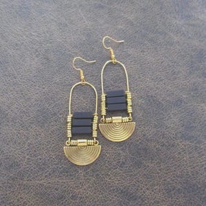 Puede incluir: Un par de pendientes dorados con un diseño geométrico. Cada pendiente presenta un gancho dorado, una sección rectangular de tres cuentas de color gris oscuro y una base semicircular con líneas concéntricas. Los pendientes se muestran sobre una superficie texturizada de color gris oscuro.