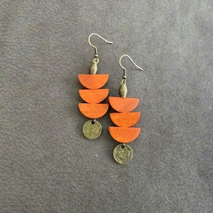 May include: A pair of dangle earrings featuring three stacked orange wooden semi-circles suspended from a bronze-toned hook. Each earring is finished with a small, round, textured bronze-toned metal disc at the bottom and a coiled metal bead.