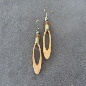May include: A pair of wooden oval earrings with brass-colored accents and ear hooks. The earrings have a light brown color with a natural wood grain. The top of each earring features a small, amber-colored bead.
