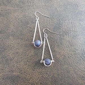 May include: A pair of silver-toned earrings with a triangular design. Each earring features a dark blue, spherical bead suspended within the triangle. The earrings have a simple, elegant design with a hook closure.