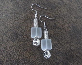 May include: A pair of silver-toned earrings. Each earring features a frosted, square bead, a textured cylindrical bead, and a small, round, textured bead. The earrings hang from a simple hook.
