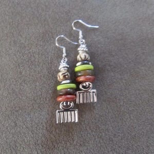 Puede incluir: Un par de pendientes de plata con cuentas de madera en tonos marrones, verdes y blancos. Los pendientes tienen un dije de peine de plata en la parte inferior.