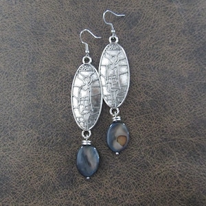 May include: Silver earrings with a detailed design of a woman carrying a basket on her head. The earrings are accented with a black and brown oval bead.