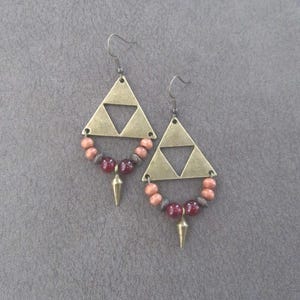 May include: A pair of brass-coloured triangle earrings with a cut-out triangle design. The earrings feature a circle of pink and burgundy beads, with a gold-coloured spike pendant. The earrings hang from a simple hook.