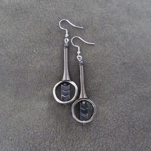 May include: A pair of drop earrings featuring a silver-toned metal coil extending from a hook, connected to a circular frame. Inside each circle, three dark gray chevron-shaped beads are stacked vertically, creating a geometric design.