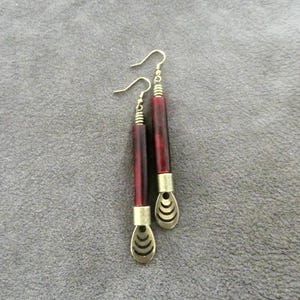 Könnte beinhalten: Ein Paar lange, rote und goldene Ohrringe mit einem dekorativen Design am unteren Ende. Die Ohrringe sind aus Holz und Metall.