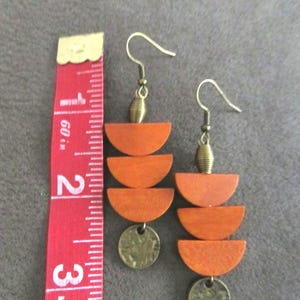 May include: A pair of dangle earrings featuring three stacked orange wooden semicircles and a small circular brass charm, suspended from brass hooks. A red measuring tape is placed alongside, showing the earrings are approximately 3 inches long.