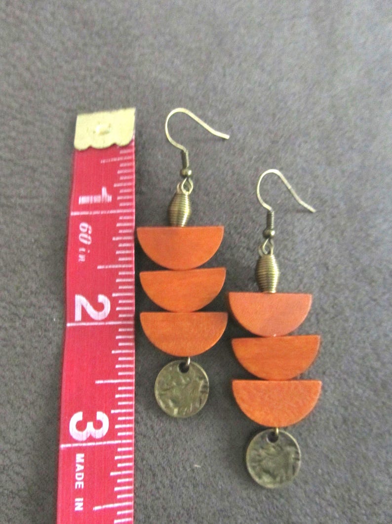 May include: A pair of dangle earrings featuring three stacked orange wooden semicircles and a small circular brass charm, suspended from brass hooks. A red measuring tape is placed alongside, showing the earrings are approximately 3 inches long.