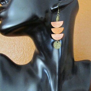 May include: A dangle earring featuring three stacked orange wooden semi-circles suspended from a bronze-toned metal hook. A small, textured, circular bronze-toned metal charm hangs from the bottom of the lowest wooden semi-circle.