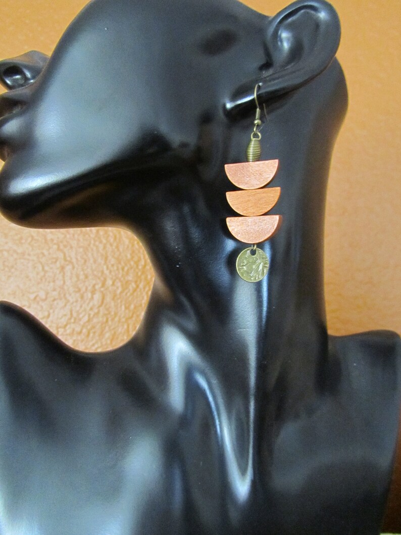 May include: A dangle earring featuring three stacked orange wooden semi-circles suspended from a bronze-toned metal hook. A small, textured, circular bronze-toned metal charm hangs from the bottom of the lowest wooden semi-circle.